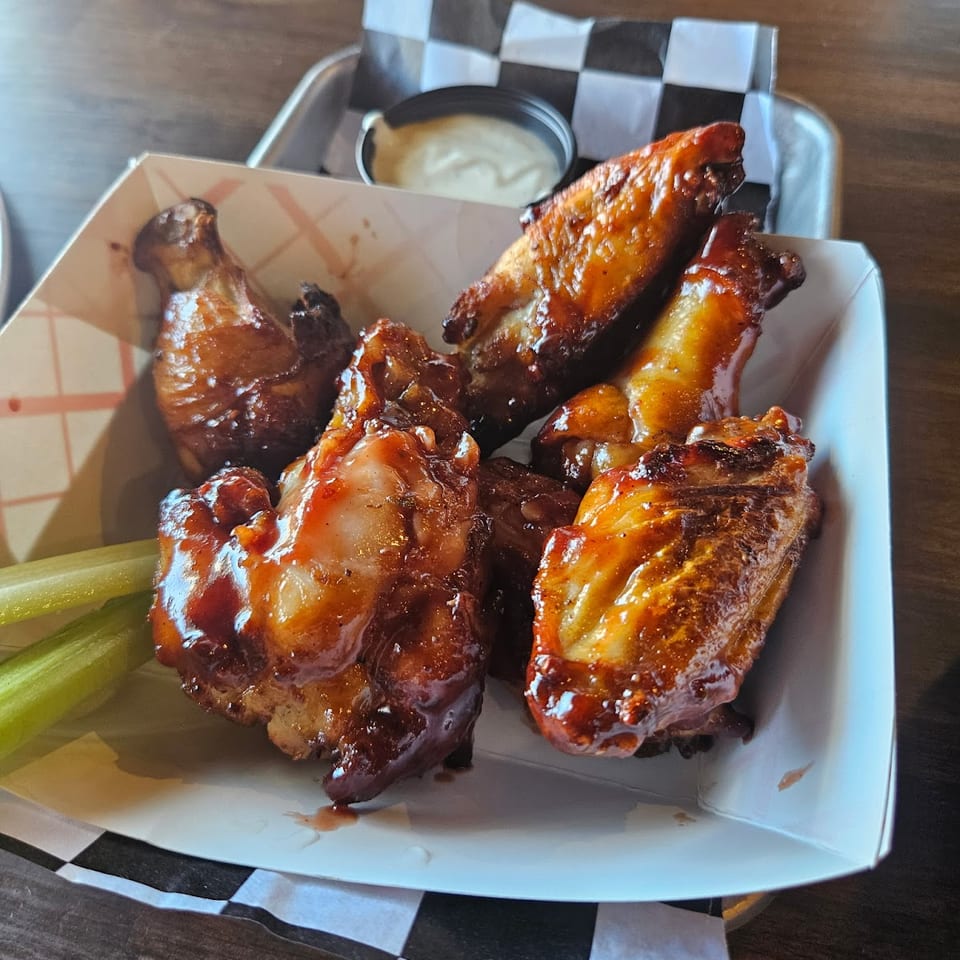 A white paper tray holds barbecue chicken wings, celery sticks, and a small cup of dipping sauce.